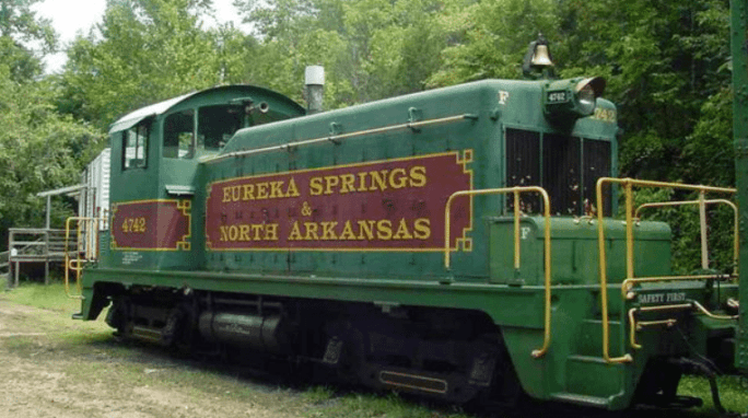 A green vintage locomotive with the lettering "Eureka Springs & North Arkansas" parked in a wooded area.