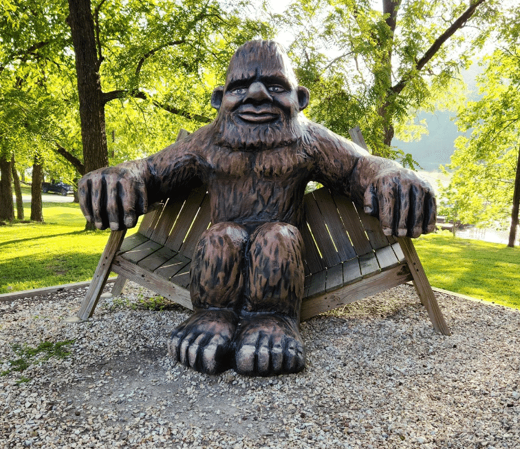 A wooden statue of Bigfoot sitting on a bench in a park.