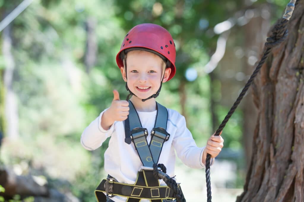 A smiling child in a red helmet gives a thumbs up while holding onto a rope outdoors.