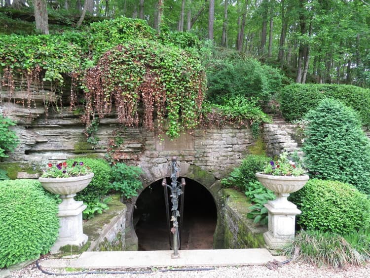 A stone archway entrance to a subterranean space, surrounded by lush greenery and decorative urns.