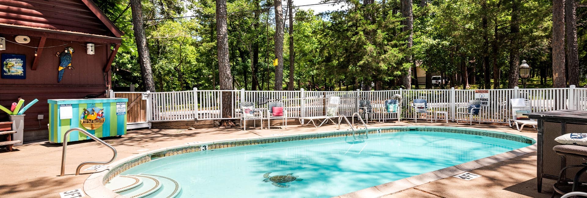 A small swimming pool surrounded by trees and lounge chairs, with a colorful poolside bar in the background.