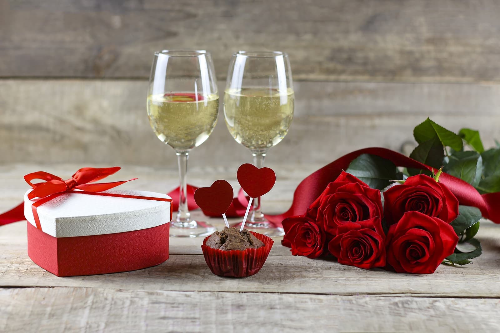 A romantic setup featuring two glasses of champagne, a red ribbon heart-shaped gift box, a chocolate cupcake with heart toppers, and a bouquet of red roses.