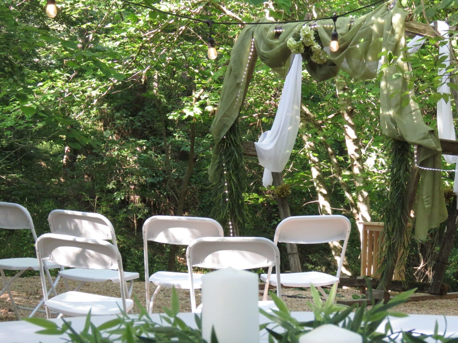 A serene outdoor wedding setup featuring white chairs, greenery, and draped fabric under soft lighting.