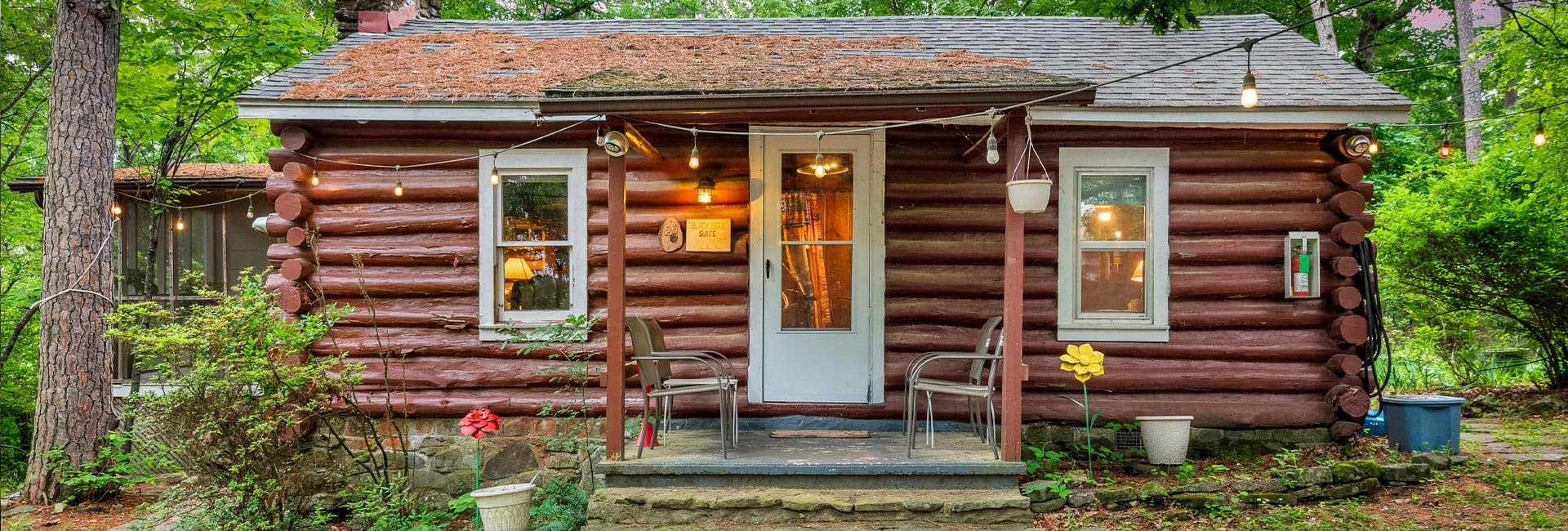A cozy log cabin surrounded by greenery, featuring a front porch with chairs and decorative plants.
