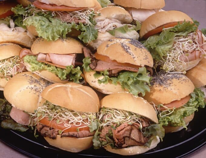 A platter piled high with assorted mini sandwiches filled with various meats, lettuce, and sprouts.