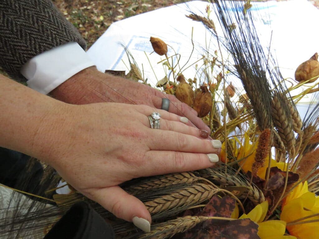 Two hands with wedding rings rest together on a rustic floral arrangement.