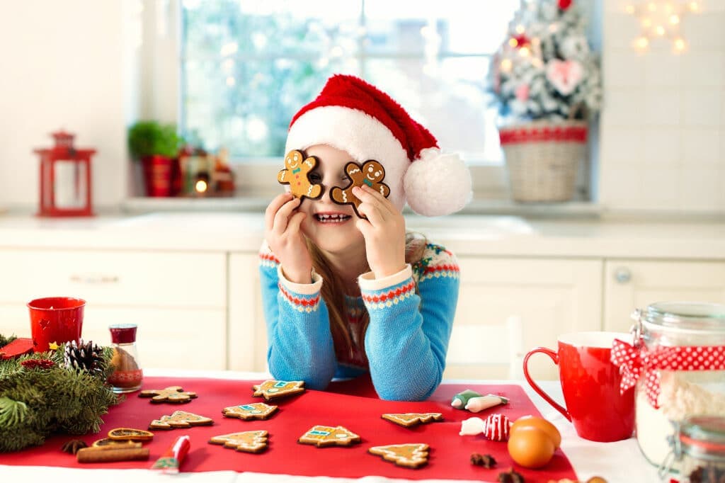 A smiling child in a Santa hat holds gingerbread cookies in front of her eyes, surrounded by festive decorations and treats on a table.