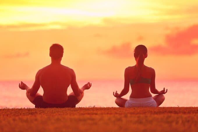 A man and woman meditate together at sunset by the ocean.