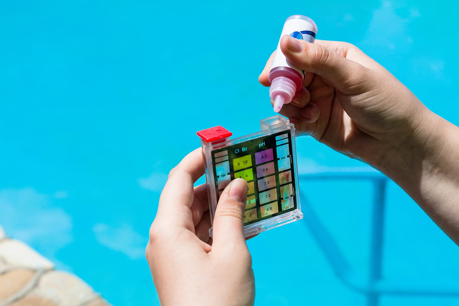 A person holds a pool water testing kit and a dropper bottle above a turquoise pool.