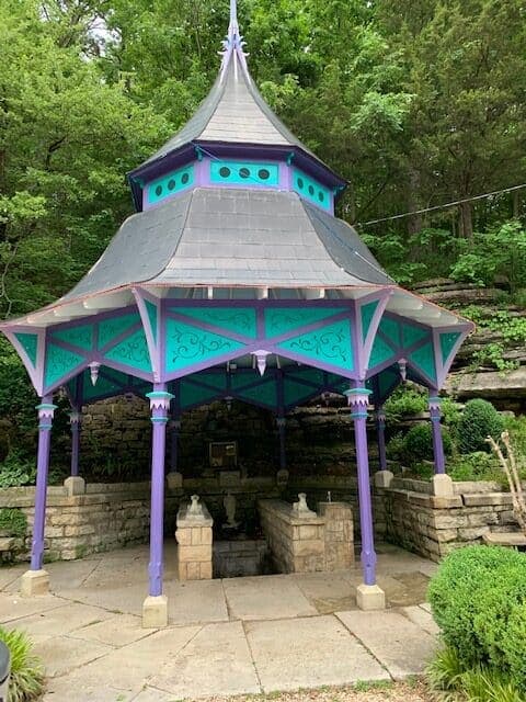 A colorful gazebo with a peaked roof situated among greenery and stone features.