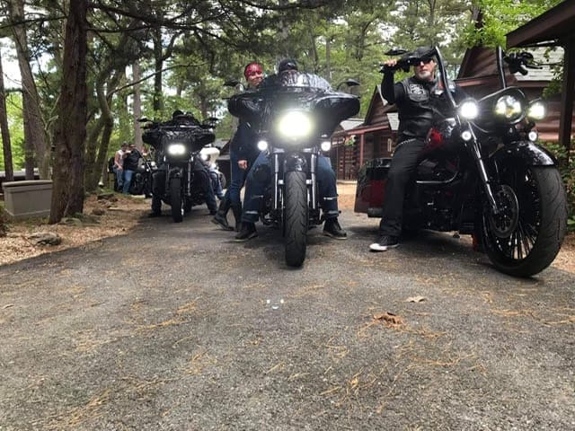 A group of motorcycles parked on a wooded path with riders standing beside them.