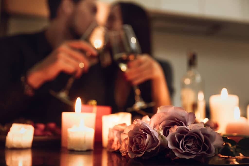 A couple toasts with glasses of wine, surrounded by candles and roses on a romantic dinner table.