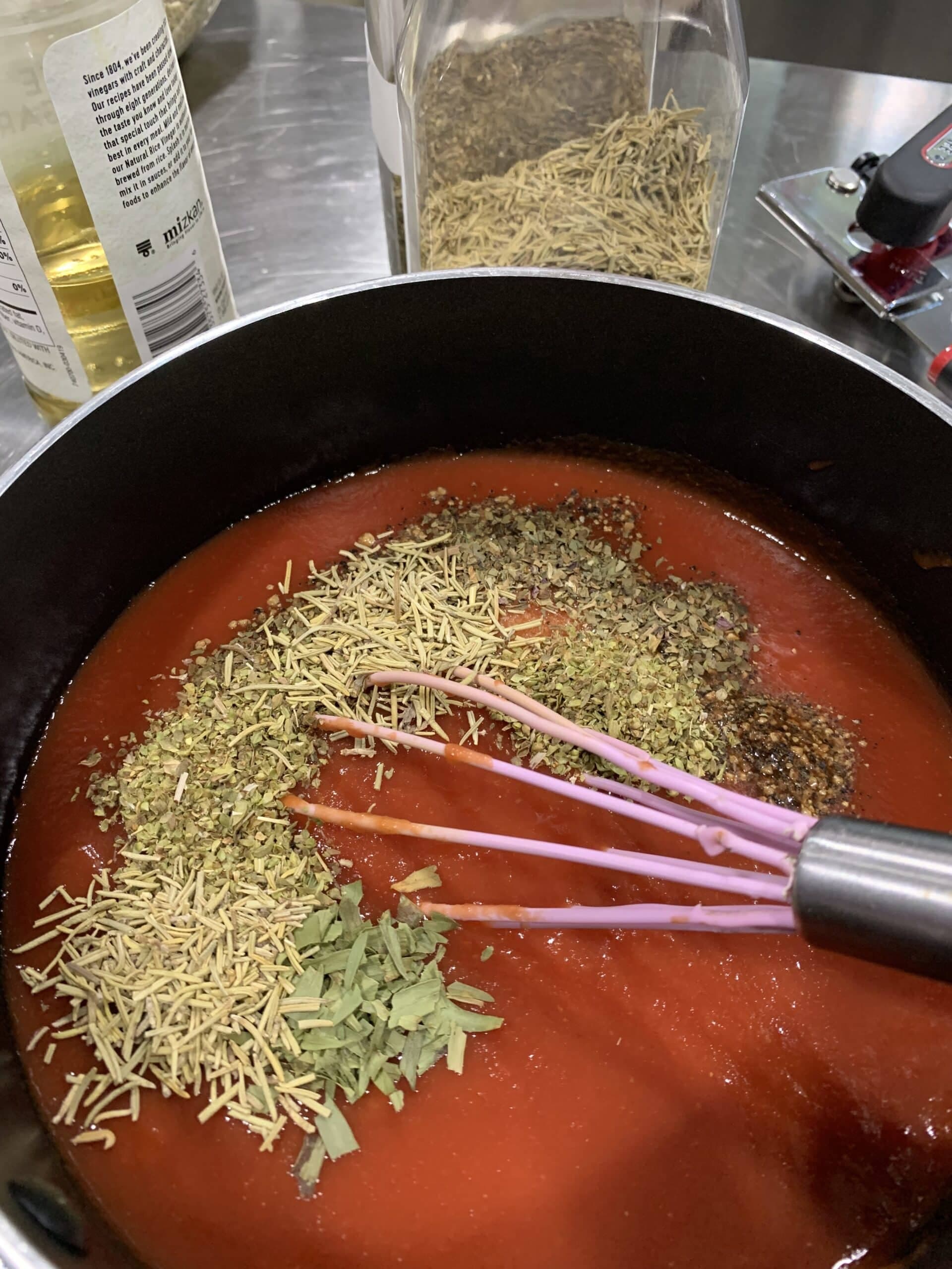 A pot of tomato sauce with various herbs and spices being mixed with a whisk.