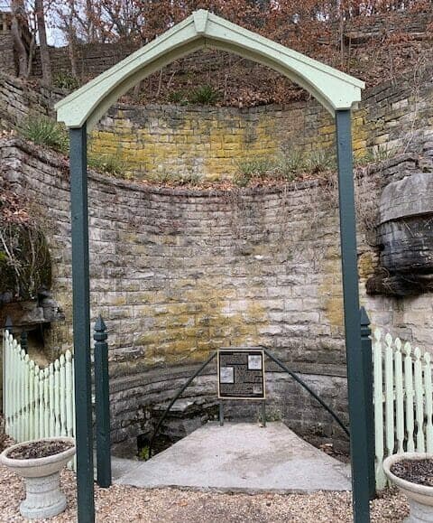 An archway frames a stone structure with a plaque, surrounded by weathered walls and greenery.