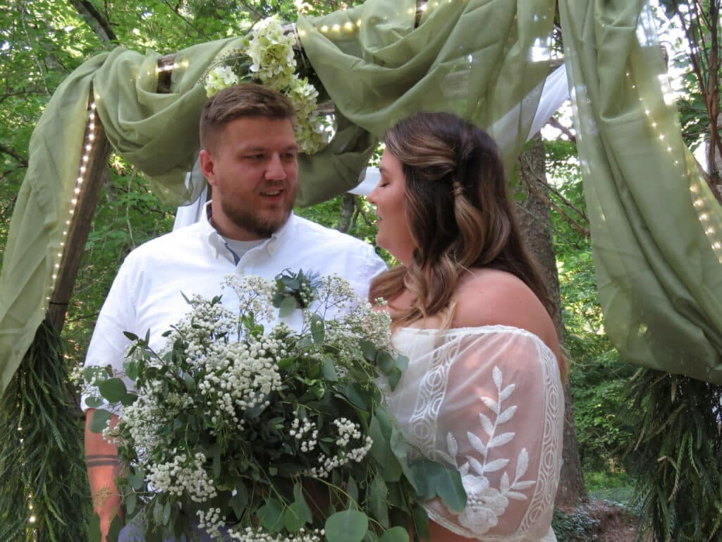 A couple gazes at each other while standing under a decorative canopy, surrounded by greenery and holding a bouquet.
