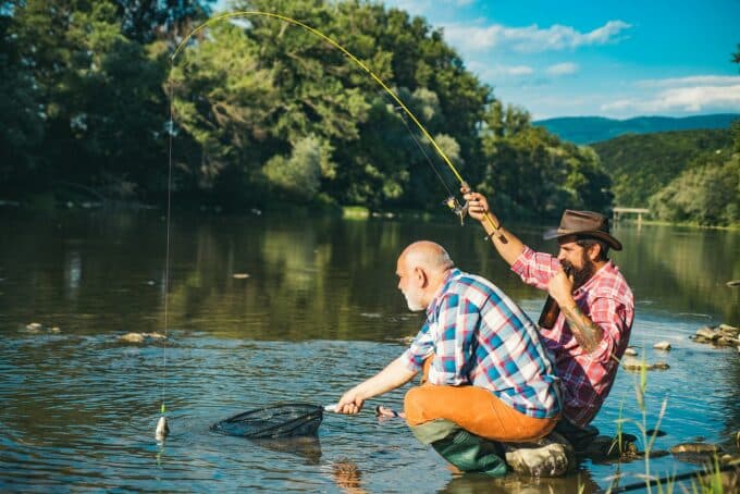 Two men are fishing by a river, one catching a fish while the other prepares a net.