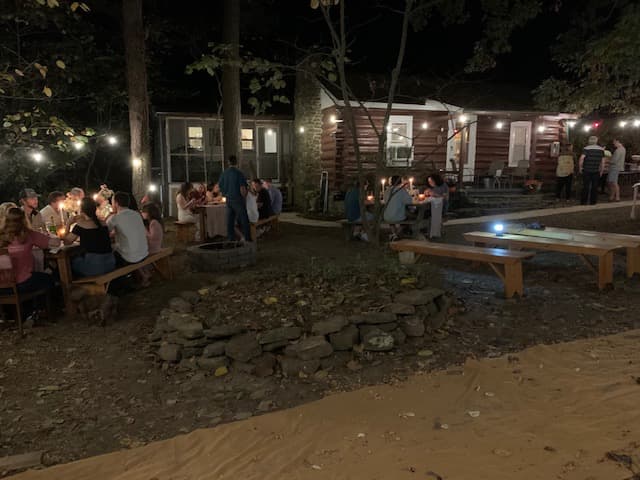 A cozy outdoor gathering at night, with people dining at tables and a log cabin illuminated in the background.