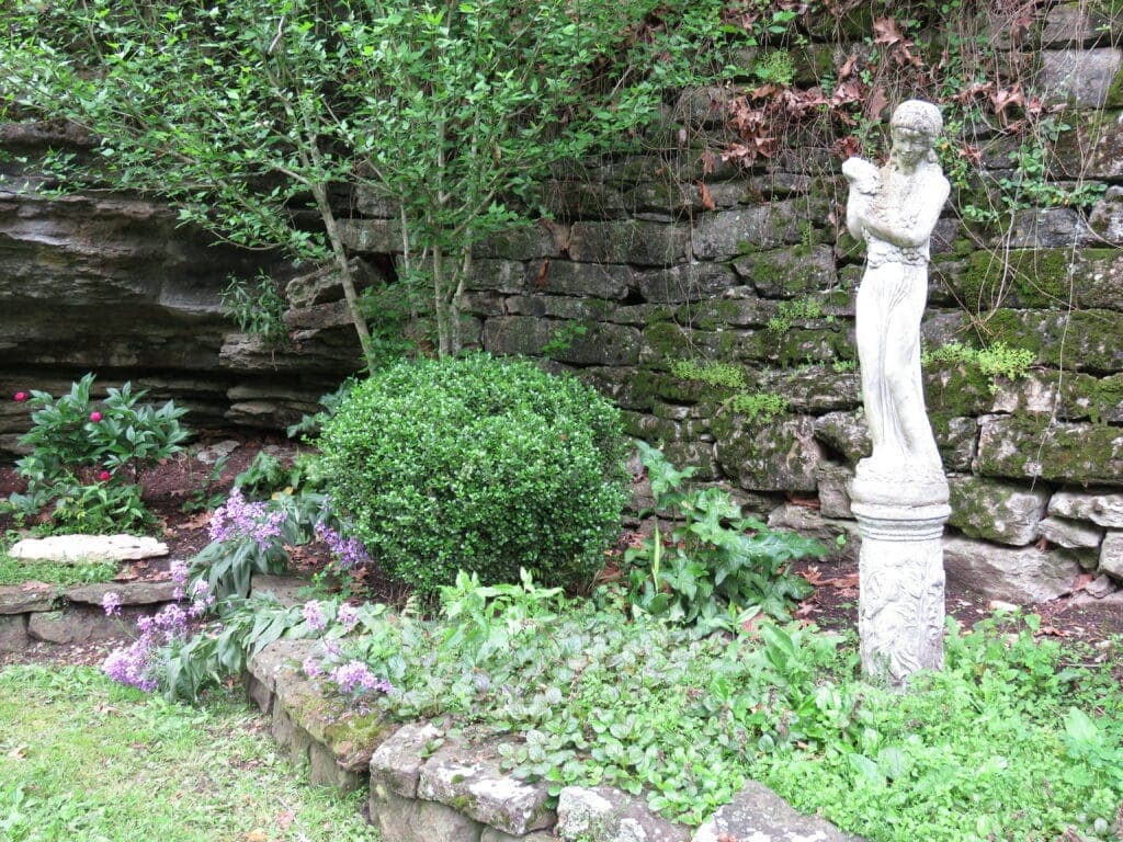 A stone statue surrounded by lush greenery and flowering plants in a garden setting.