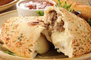 A plate with a stuffed pastry, partially broken open to reveal meat and cheese filling, accompanied by a small bowl of salsa.