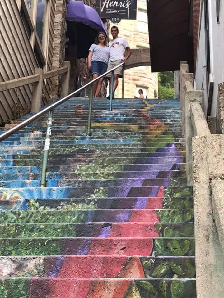 A couple stands on colorful, mural-covered stairs, with buildings and a sign visible in the background.