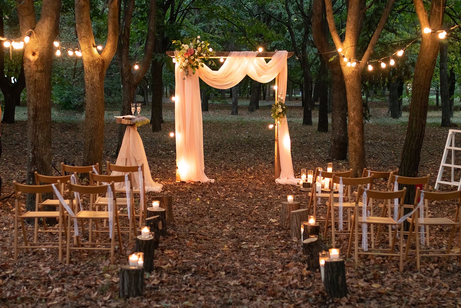 A beautifully decorated outdoor wedding venue with a floral arch, intimate seating, and soft lighting amidst trees and autumn leaves.