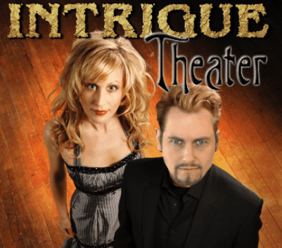A man in a black suit and a woman in a silver dress pose in front of the text "Intrigue Theater" on a wooden stage.
