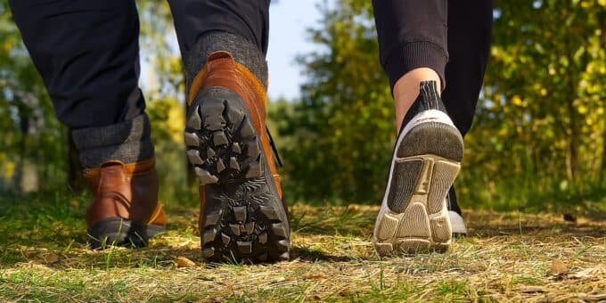 Two pairs of feet walking on a grassy path, one in hiking boots and the other in athletic shoes.