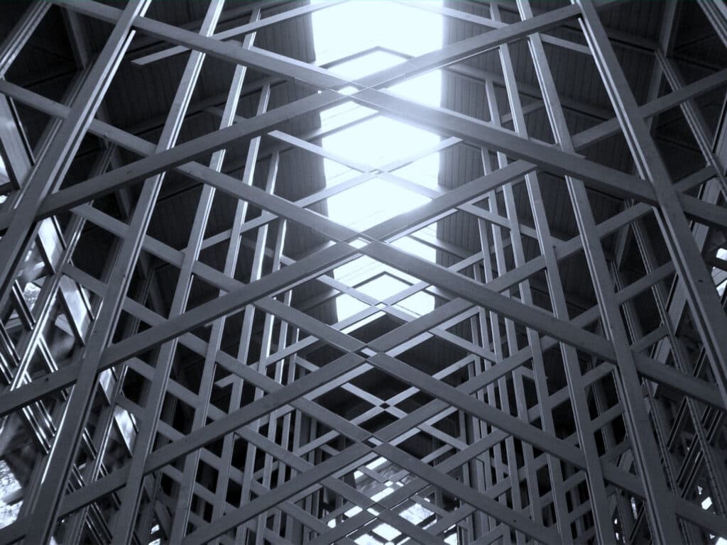 Black and white image of an intricate lattice of metal beams converging towards a bright skylight.