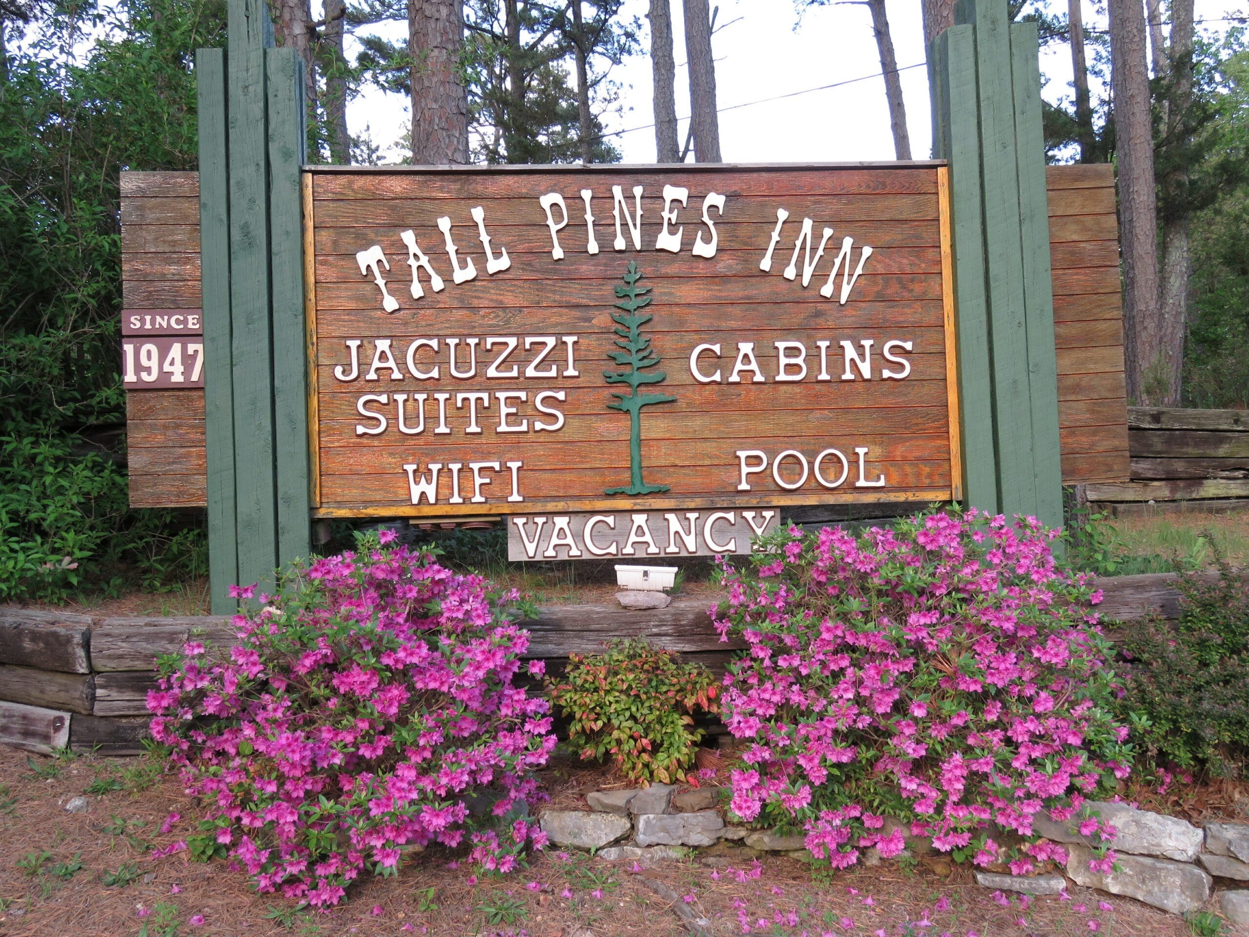 Sign for Tall Pines Inn featuring amenities like jacuzzi suites and pool, surrounded by blooming flowers.