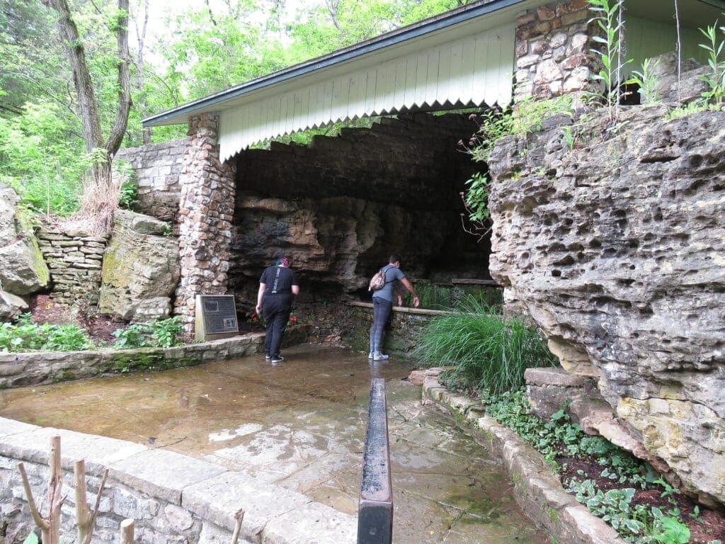 Two people explore a rocky cave entrance surrounded by greenery.