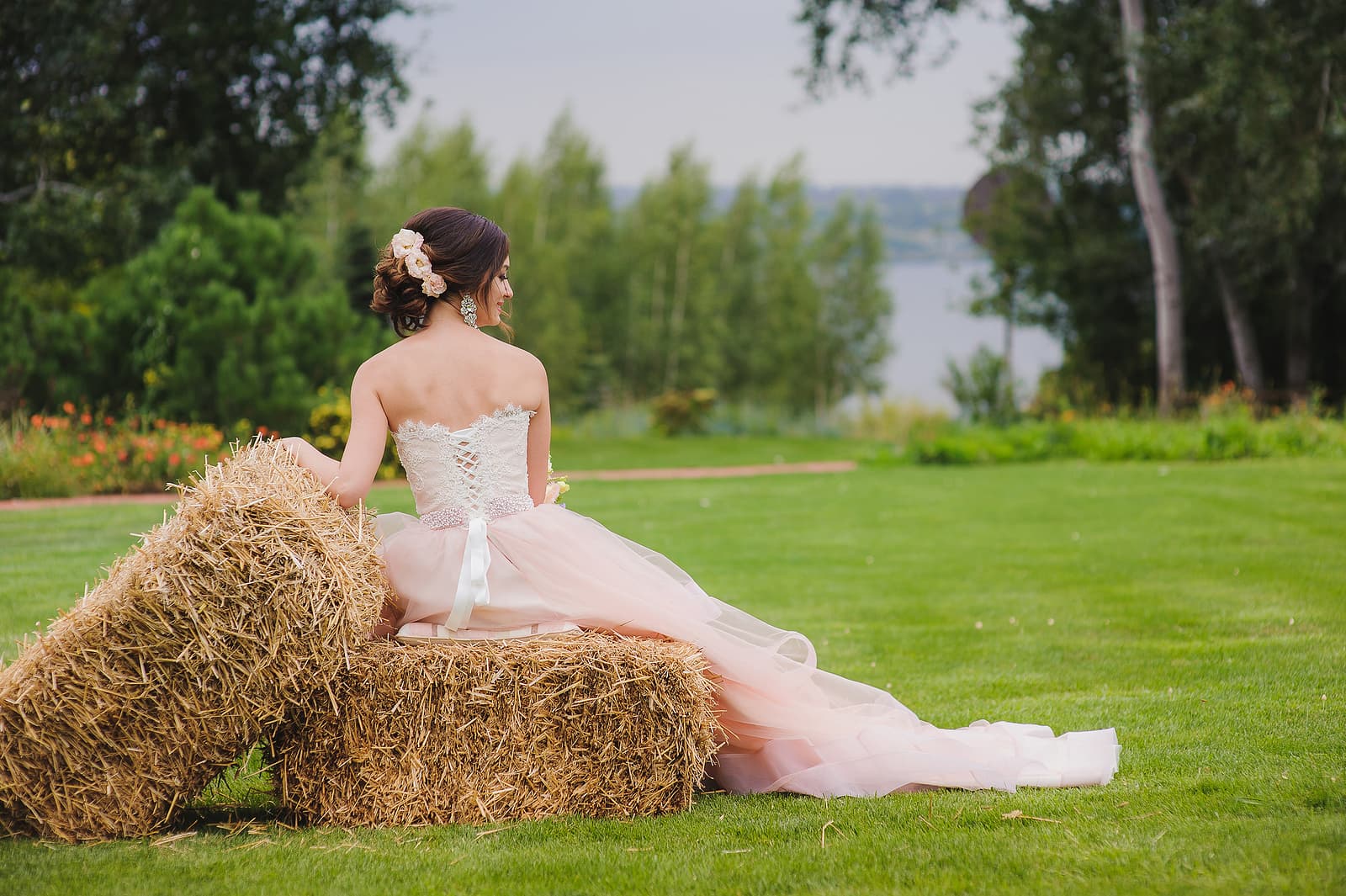 A bride in a flowing pink gown sits on hay bales, gazing at a scenic landscape.