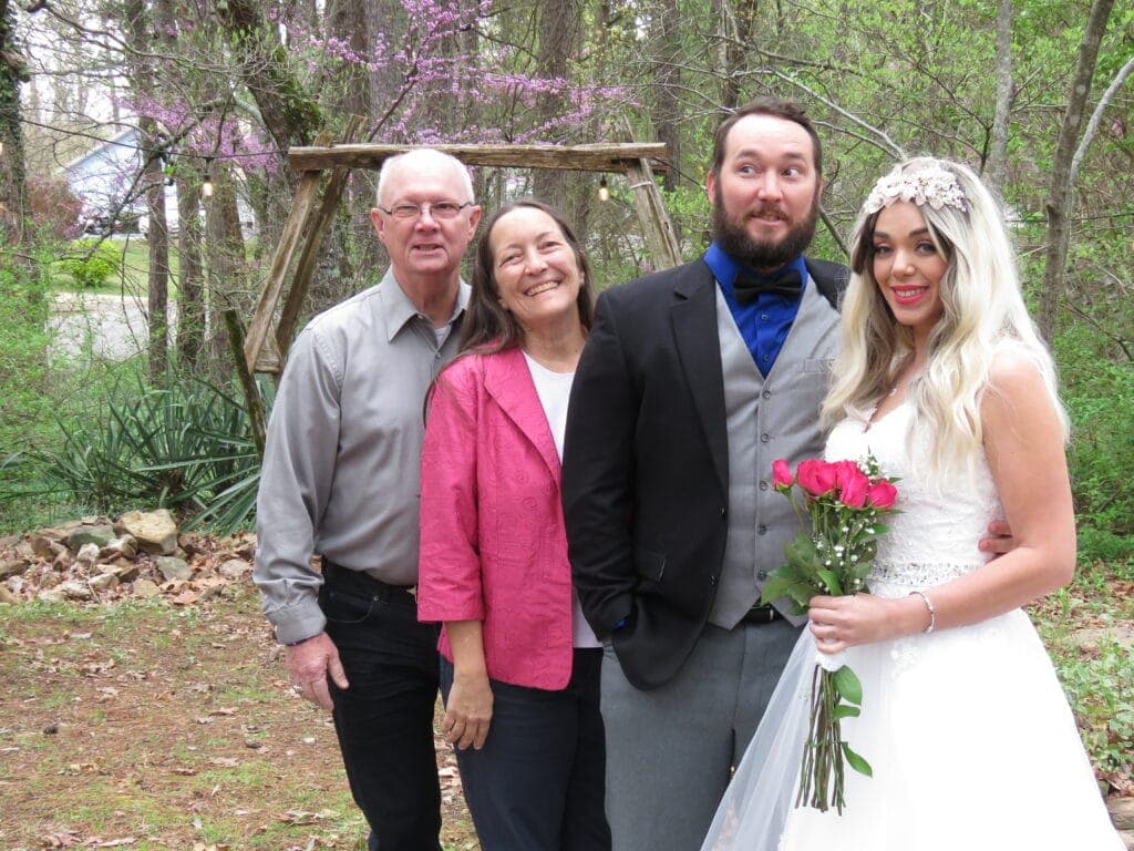 A bride in a white gown holds a bouquet while posing with three smiling family members outdoors.