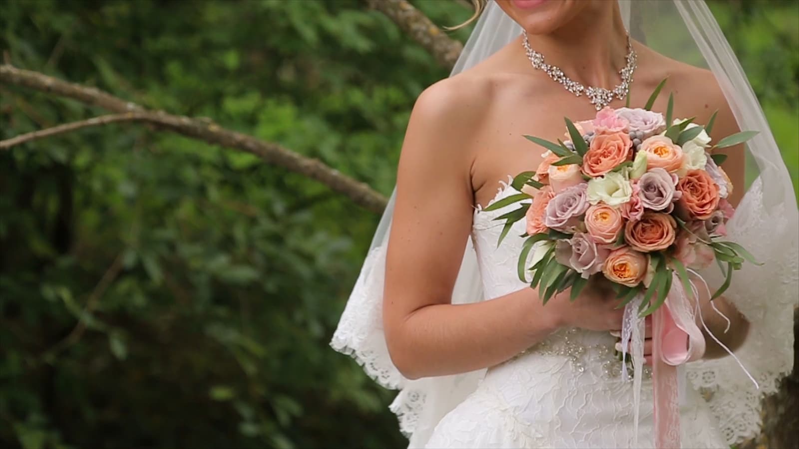 A bride in a white wedding dress holds a bouquet of pastel roses in a lush, green setting.