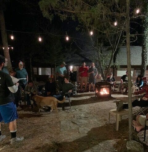 A group of people gathers around a fire pit in a wooded area at night, with string lights overhead.
