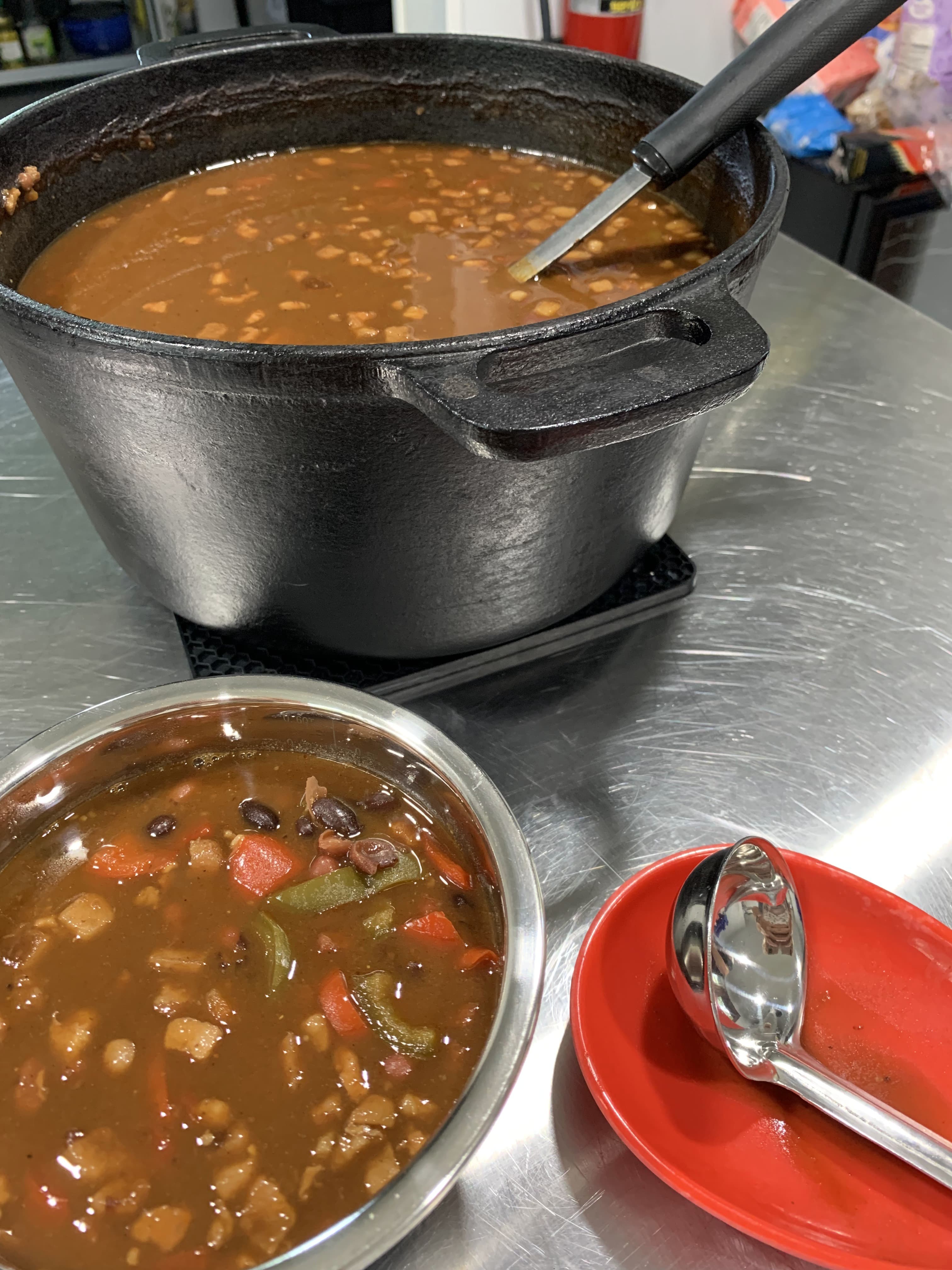 A large black pot of soup sits next to a small bowl of soup and a spoon on a metal countertop.
