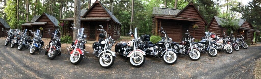 A lineup of various motorcycles parked in front of wooden cabins in a wooded area.