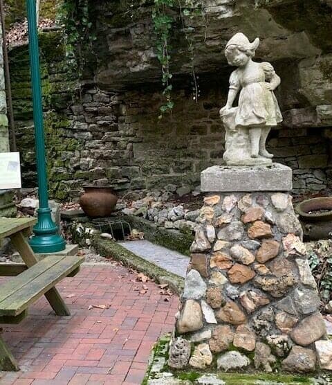 A small stone statue of a girl stands on a rocky pedestal in a garden setting with a wooden bench and a rustic backdrop.