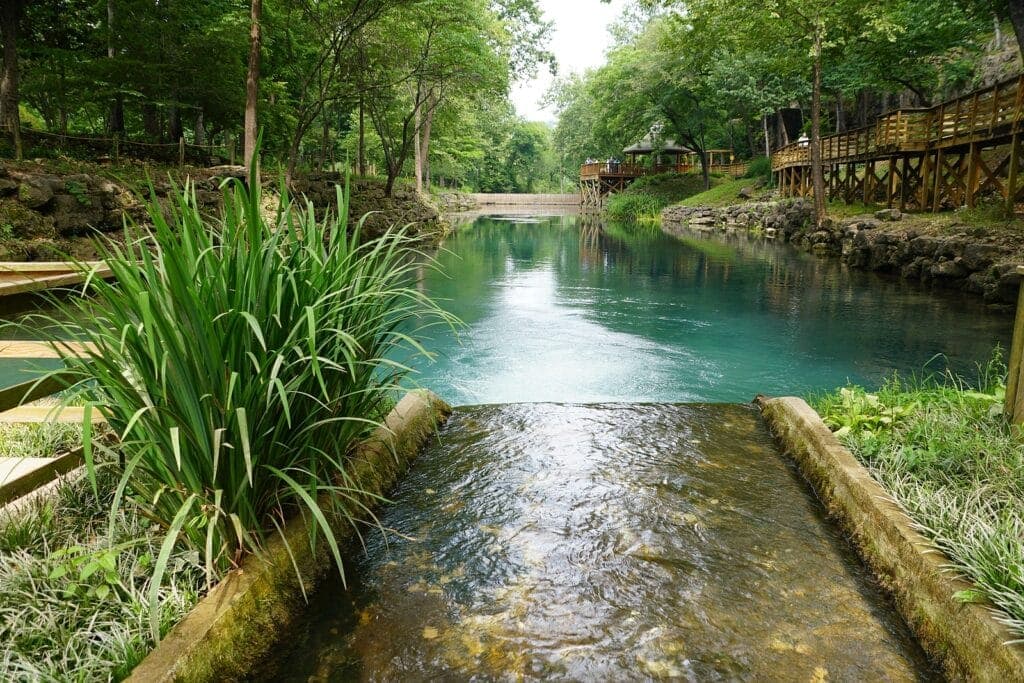 A tranquil stream flows into a clear, blue-green body of water surrounded by lush greenery and wooden walkways.