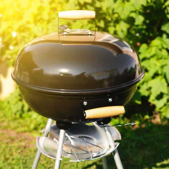 A black charcoal grill with a wooden handle, positioned on a stand in a sunny garden.