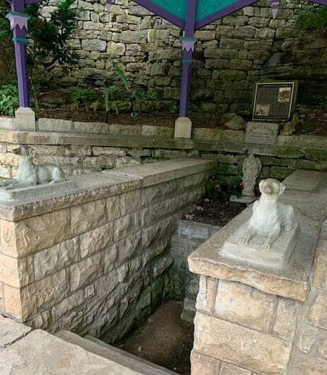 A stone alcove featuring two sculptures of dogs and a plaque, surrounded by natural stone walls and greenery.