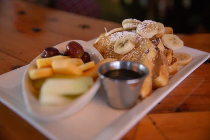 A plate of French toast topped with sliced bananas, powdered sugar, and syrup, accompanied by a fruit medley of grapes, cantaloupe, and honeydew.
