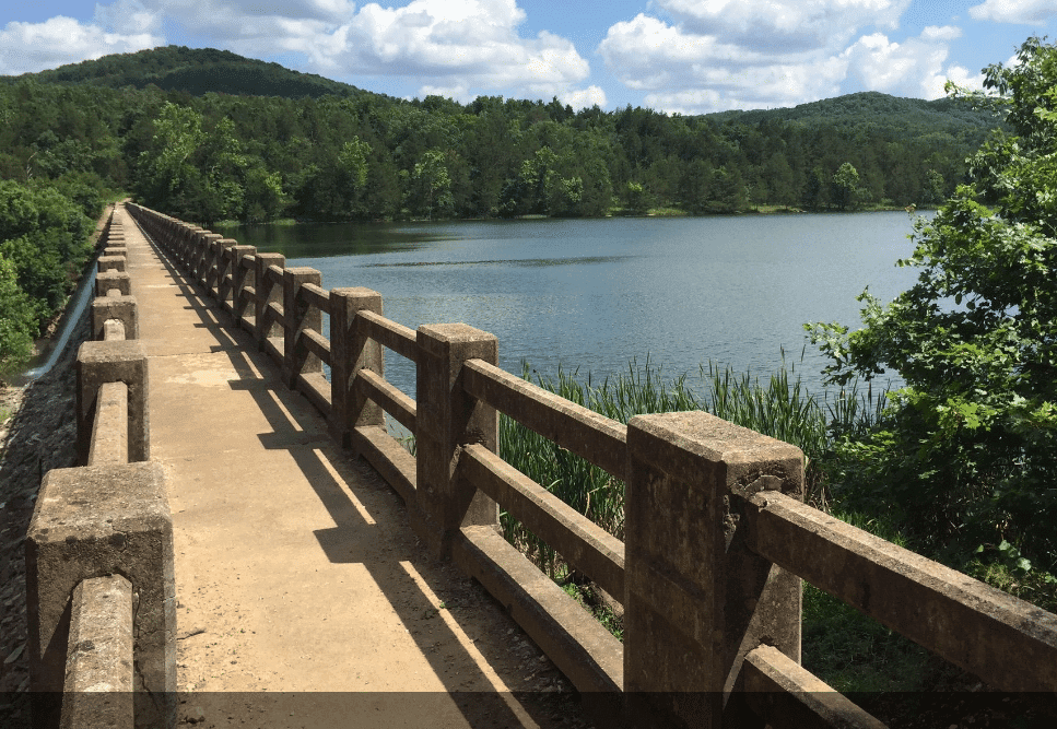 A concrete path with a railing stretches over a calm lake surrounded by greenery and hills under a partly cloudy sky.