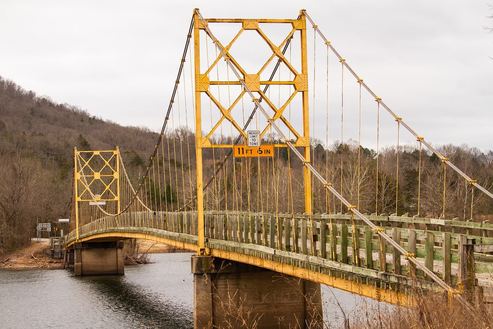 A yellow suspension bridge spans a river surrounded by bare trees under a cloudy sky.