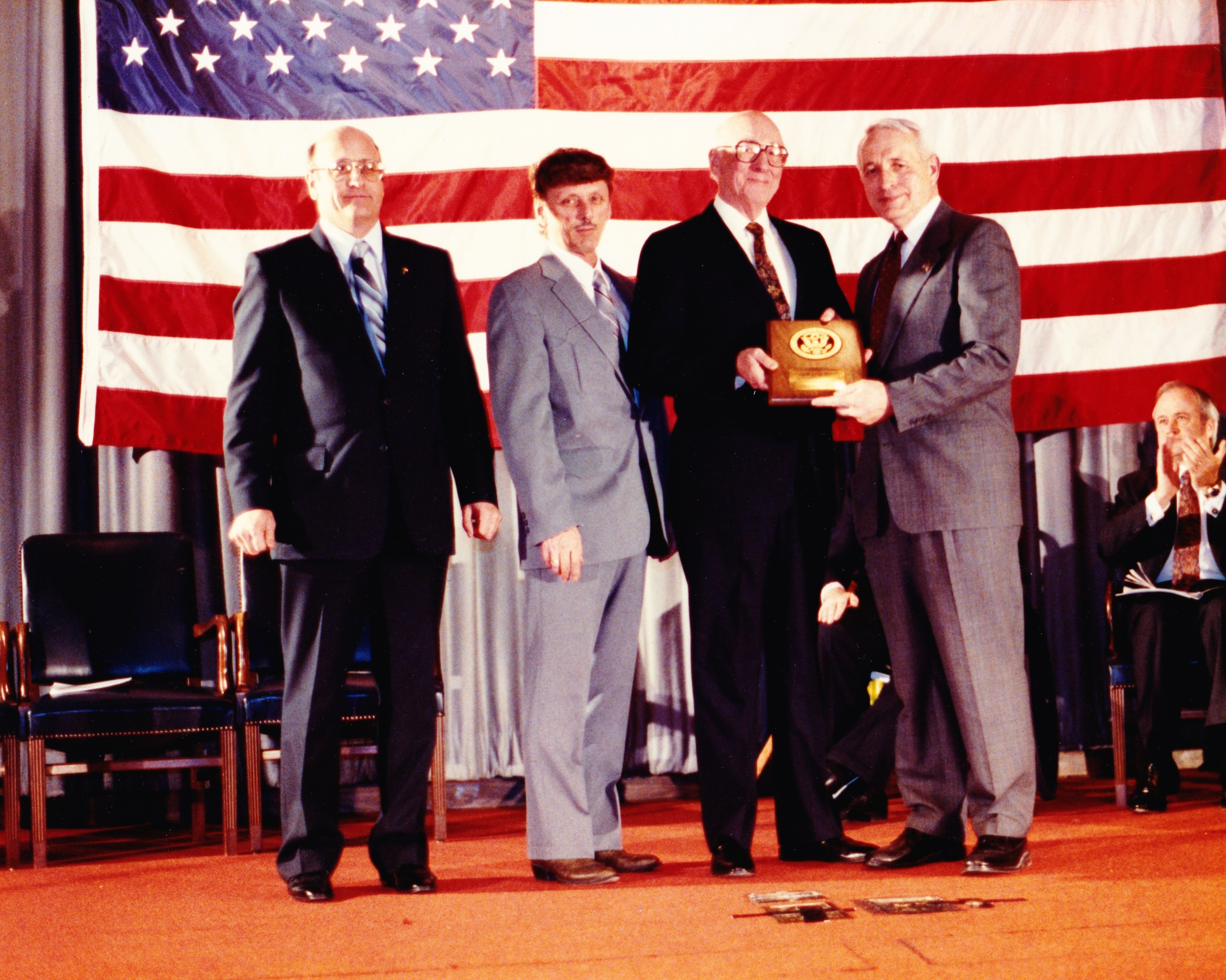 three men receiving an award from another man with the american flag hanging behind them