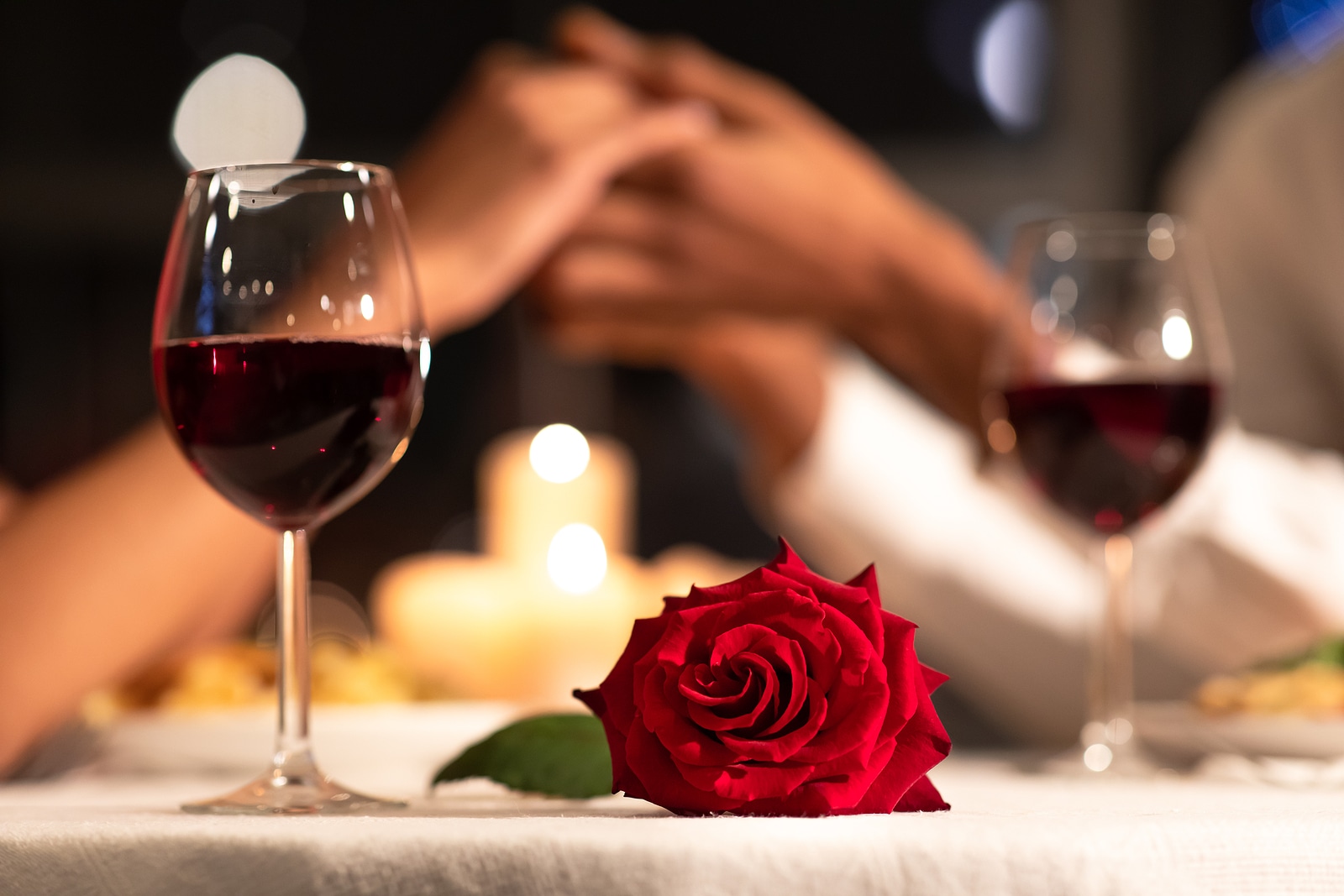 two wine glasses with a rose in the middle and two people holding hands