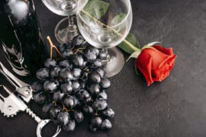 Purple grapes sitting between two wine empty wine glasses and one long stem red rose sitting next to the wine glasses