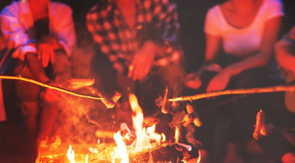 group of friends sitting around a campfire at night roasting marshmallows on sticks