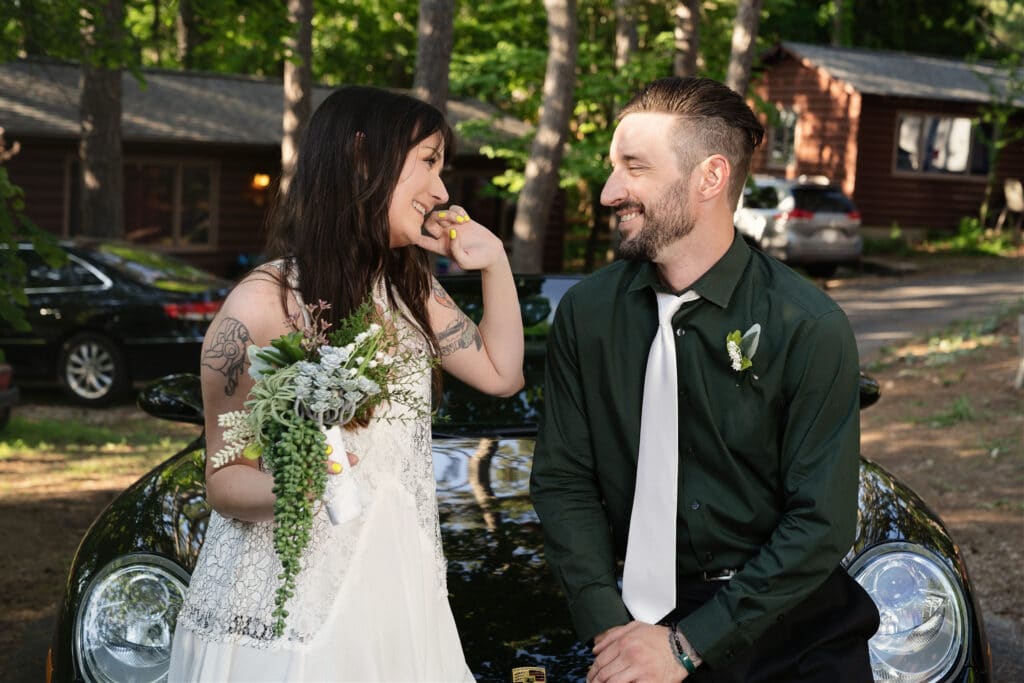 bride and groom sitting on hood of car smiling at each other cabins in the back ground