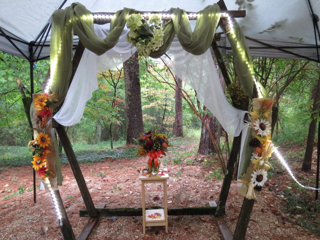 Rustic wedding arch decorated in white and green drapes and green flowers and lights with a fall bouquet set in the middle
