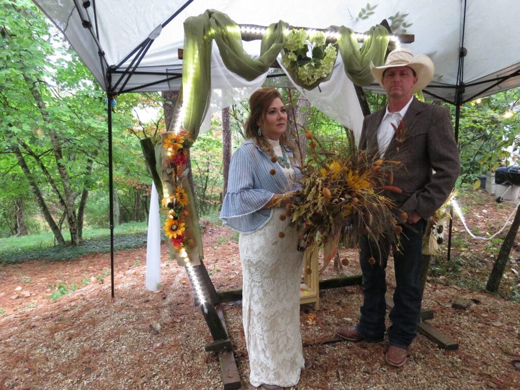 man and woman standing in front of a decorated rustic wedding arch in colors of green and white and flowers the bride is holding a large bouquet of fall foliage